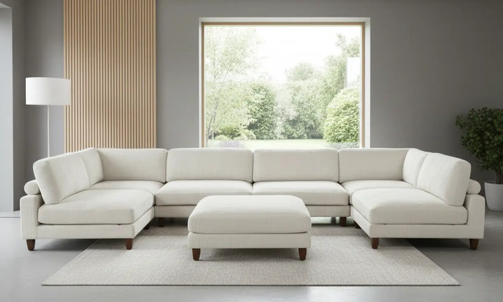 Beige sectional sofa with ottoman in a room with large windows and a plant.