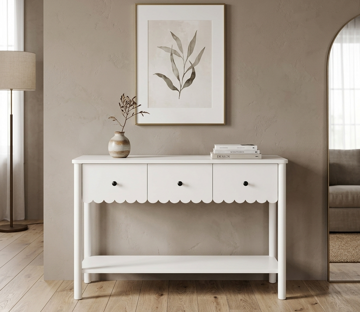 White console table with scalloped edge in a room with a lamp, plant, and framed picture on the wall.